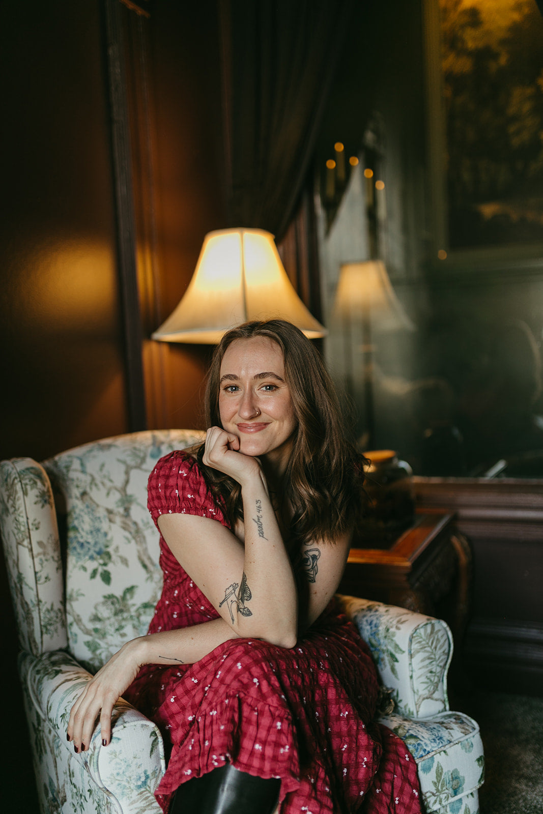 Woman in a red dress sitting on a floral armchair in a warmly lit room.