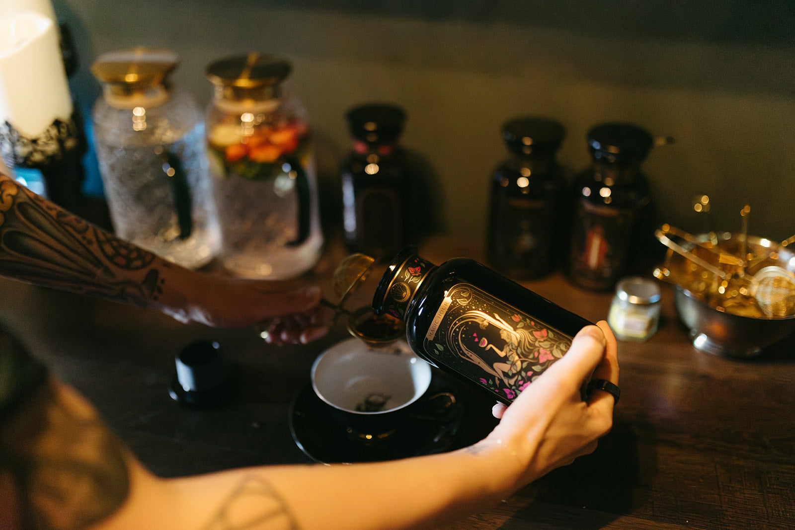 Person pouring a dark liquid from a bottle into a small cup on a wooden surface with various bottles in the background.